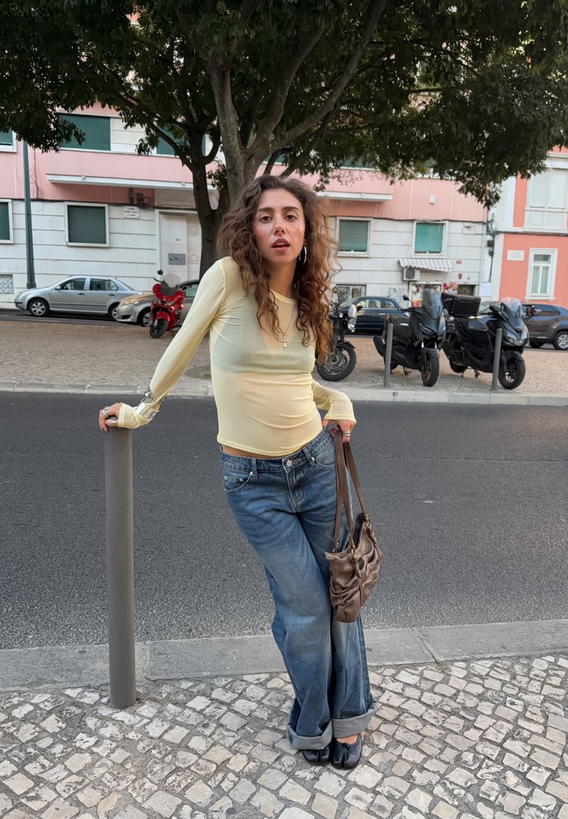 Jeune femme aux cheveux bouclés appuyée contre un poteau dans une rue de la ville, portant un haut léger à manches longues, un jean large et tenant un sac à main marron.