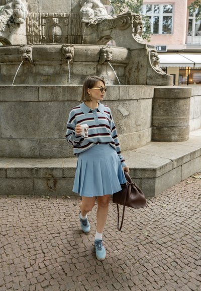 Polo a rayas en azul y marrón, falda azul claro con pliegues y zapatillas azules. Bolso marrón y taza de café. Fuente de piedra al fondo.