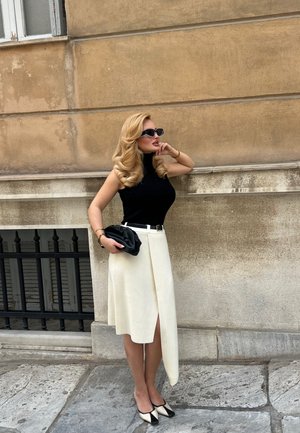 Mujer con cabello largo y rubio que lleva una blusa negra sin mangas, falda blanca asimétrica, tacones blancos y negros y gafas de sol, de pie contra una pared beige.