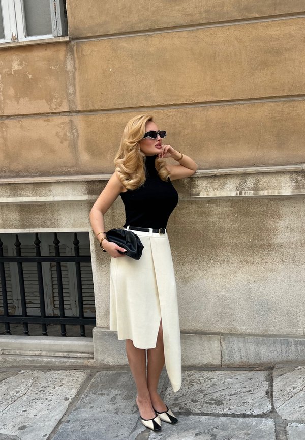 Femme aux longs cheveux blonds portant un haut noir sans manches, une jupe asymétrique blanche, des talons noirs et blancs et des lunettes de soleil, se tient contre un mur beige.