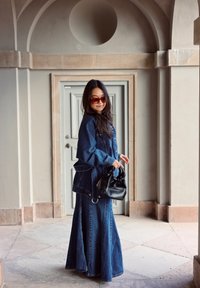 Denim jacket and skirt in dark blue, long skirt with flared hem. Accessories include a black handbag and oversized brown sunglasses.