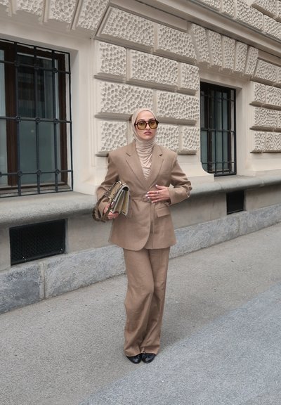 Traje marrón claro a medida con una chaqueta estructurada y pantalones de pierna ancha. Combinado con un hiyab beige y gafas de sol grandes, sosteniendo un bolso estampado.