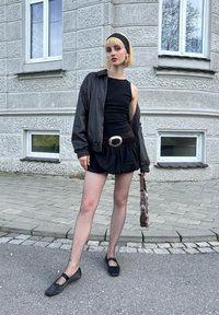 Μαύρο αμάνικο φόρεμα με πλισέ φούστα, σε συνδυασμό με μαύρο δερμάτινο τζάκετ bomber, καλσόν ψαθωτής ύφανσης και μαύρες επίπεδες παπούτσια. Καφέ ζώνη με μεγάλη ασημένια αγκράφα και τσάντα χειρός.