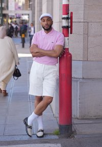 Polo shirt rosa, pantaloni corti bianchi con puffs, mocassini neri e beige e calze bianche. Appoggiato a un palo rosso e bianco su una strada.