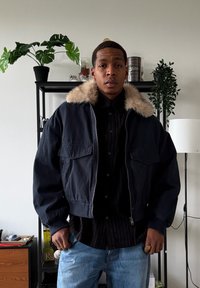 Blouson aviateur bleu foncé raccourci avec un col en fausse fourrure, doté de deux poches avant et d'une fermeture éclair, porté sur une chemise noire boutonnée.