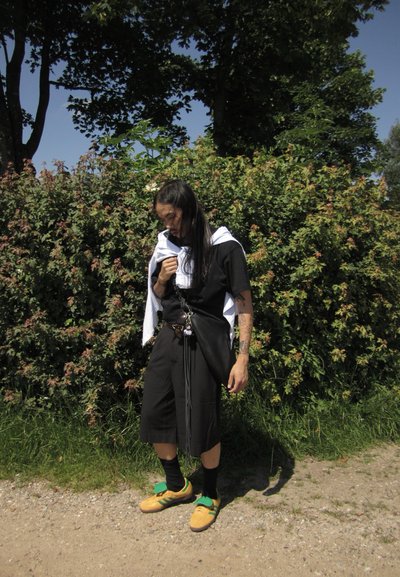 Camisa negra de corte holgado con mangas cortas, pantalones cortos casuales, zapatillas verdes y amarillas, toalla blanca colgada del hombro, de pie junto a vegetación.