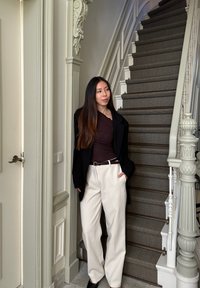 Fekete zakó egy barna felsőn, krémszínű, széles lábú nadrág övvel, áll egy lépcsőház mellett, melynek kidolgozott fehér korlátja és szürke szőnyege van.