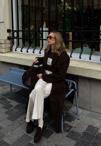 Hnědý kabát, polo tričko s černobílými pruhy, krémové kalhoty s širokými nohavicemi, čokoládové mokasíny, nese hnědou kabelku, sedí na modré lavici.