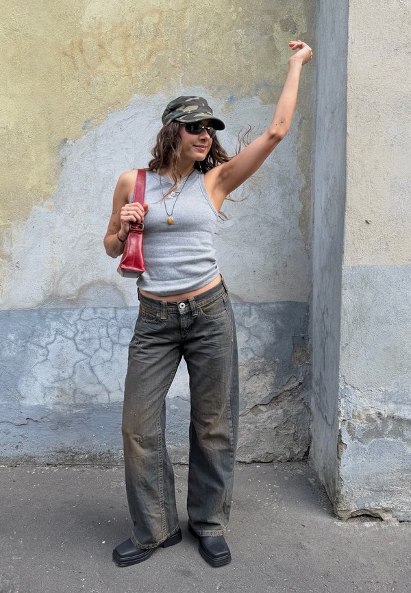 Personne portant un débardeur gris, un jean foncé, une casquette camouflage, des lunettes de soleil et des chaussures noires, tenant un sac rouge avec un bras levé contre un mur texturé.