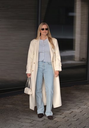 Mujer que lleva un abrigo largo color crema, blusa azul claro con botones, vaqueros azul claro, zapatos oscuros, gafas de sol y sostiene un pequeño bolso crema.