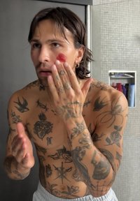 Tatowierter, oberkörperfreier Mann mit dunklem Haar trägt rote Creme auf seine Wangen in einem Badezimmer mit gefliester Wand und einem Regal voller Toilettenartikel auf.