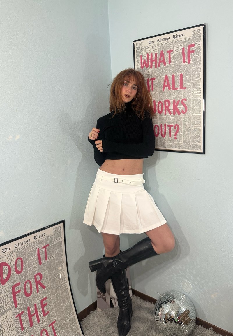 Giovane donna con dolcevita nera, gonna bianca e stivali neri posa in un angolo con poster motivazionali e una palla da discoteca sul pavimento.