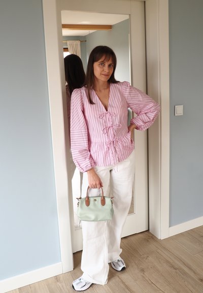 Mujer con una blusa de rayas rosa, pantalones blancos y zapatillas, de pie sosteniendo un pequeño bolso verde en una puerta con un espejo detrás de ella.