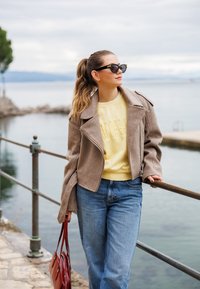 Veste en laine marron avec grands revers, sweatshirt jaune avec texte en relief, jeans bleu clair et sac à main rouge, au bord de l'eau.