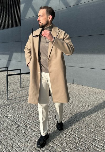 Abrigo beige, suéter de cuello alto gris, pantalones blancos y zapatos negros. El abrigo tiene un corte recto; gafas de sol sujetas en una mano. Pavimento texturizado de fondo.