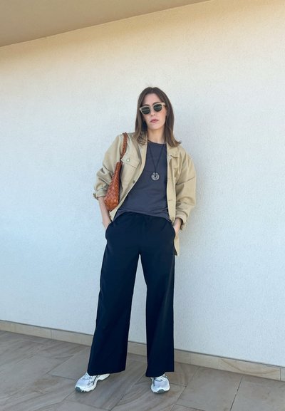 Chaqueta beige de gran tamaño, camisa gris, pantalones de pierna ancha negros, zapatillas plateadas y bolso de hombro trenzado marrón; las gafas de sol realzan el conjunto.