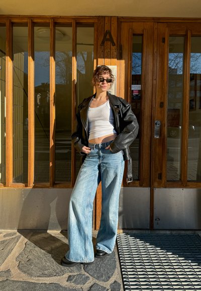 Chaqueta de cuero negra de moto, top corto blanco, pantalones vaqueros holgados de mezclilla ligera en azul, y botas de plataforma negras, con puertas de madera de fondo.