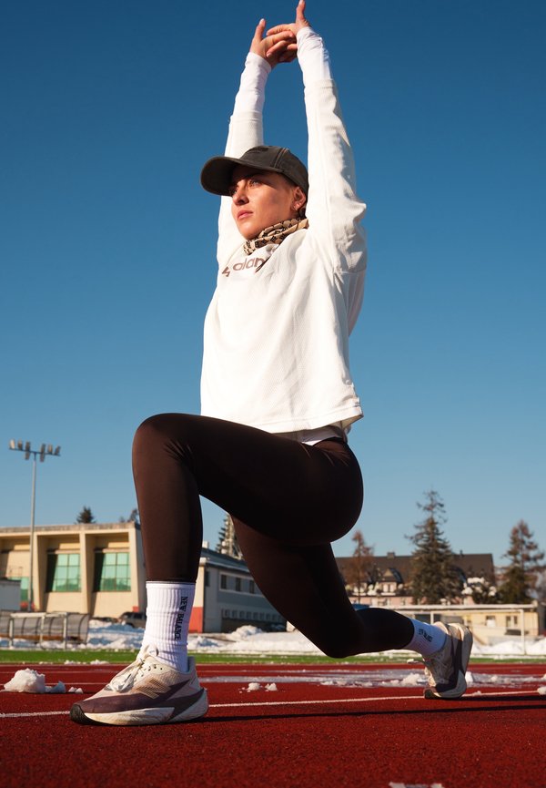 Mujer con ropa deportiva estirando una pierna hacia adelante con los brazos levantados en una pista roja al aire libre bajo un cielo azul despejado.