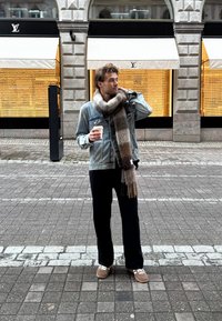Veste en denim, écharpe grise à motif en fourrure, pantalon noir, baskets brunes avec accents blancs. Tient une tasse de café, se tient sur un pavé en pierre.