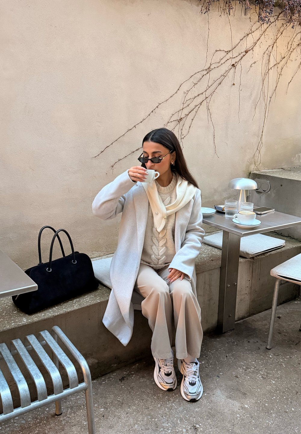 Manteau gris clair sur un pull beige en maille torsadée, écharpe crème, pantalon beige et baskets blanches. Sac à main noir posé sur un banc en pierre.