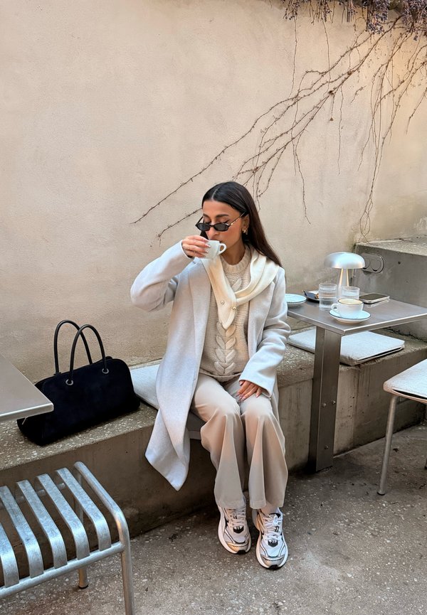 Lichtgrijze jas over een beige kabelgebreide trui, crèmekleurige sjaal, beige broek en witte sneakers. Zwarte handtas op een stenen bank.