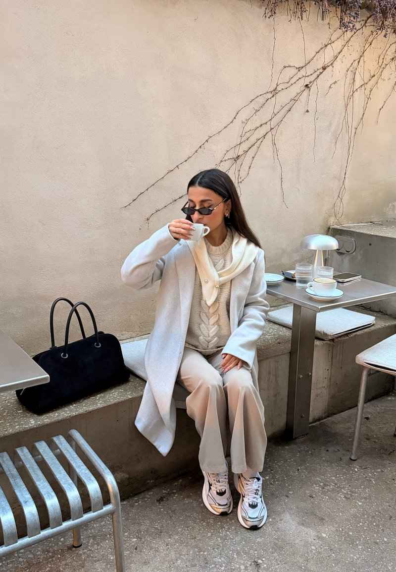 Manteau gris clair sur un pull beige en maille torsadée, écharpe crème, pantalon beige et baskets blanches. Sac à main noir posé sur un banc en pierre.