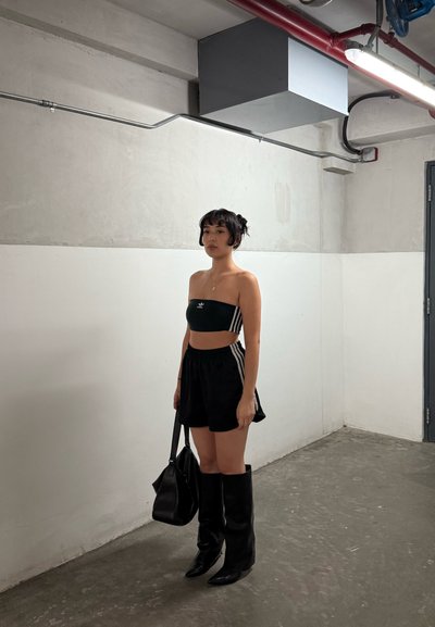 Mujer joven vestida con un top tubo negro de Adidas y shorts, con botas negras, sosteniendo un bolso negro, de pie en una habitación industrial minimalista.