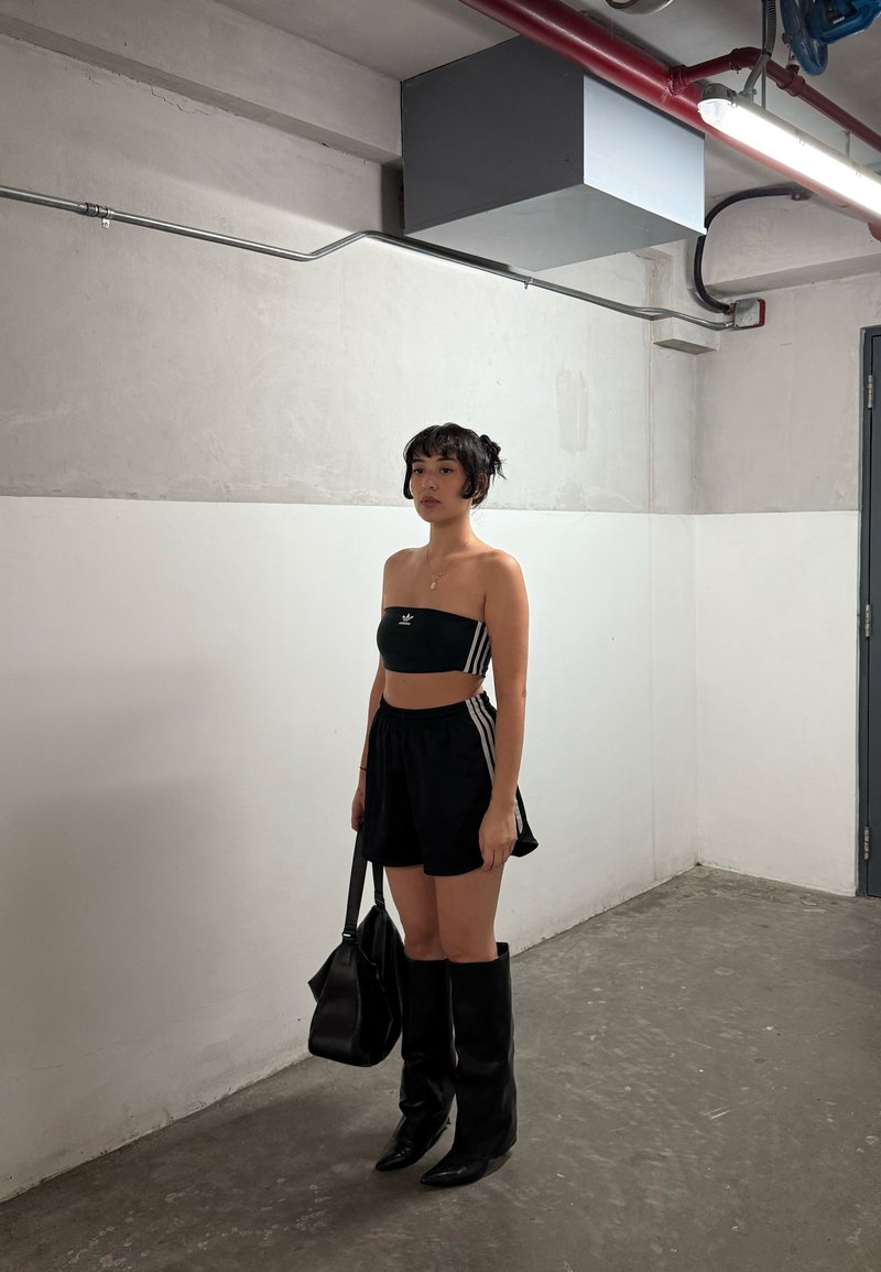 Jovem mulher vestindo um top preto da Adidas e calções com botas pretas, segurando uma bolsa preta, a pôr-se de pé numa sala industrial minimalista.