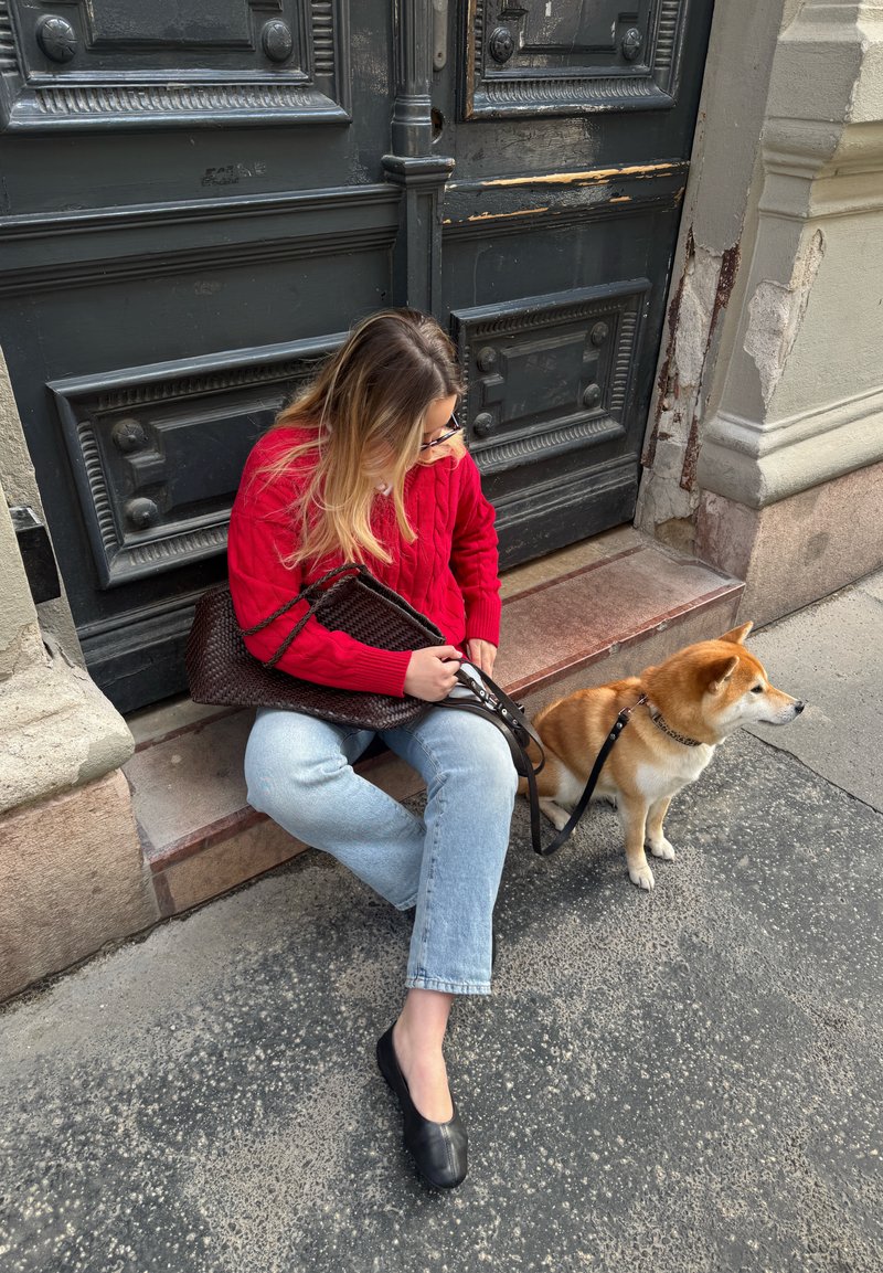 Femme en pull rouge assise sur une marche à côté d'une porte sombre ornée, tenant une laisse attachée à un petit chien fauve regardant vers la droite sur un trottoir en ville.