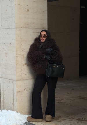 Mujer con un abrigo marrón esponjoso, pantalones negros, guantes, bufanda y gafas de sol, sosteniendo un bolso negro, parada junto a un pilar de piedra con nieve cerca.