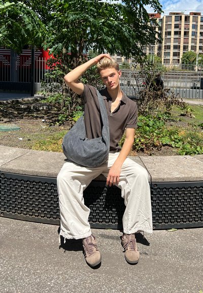 Joven con camisa marrón y pantalones blancos sentado en un banco de piedra curvado al aire libre, sosteniendo una bolsa gris y tocándose el cabello rubio.