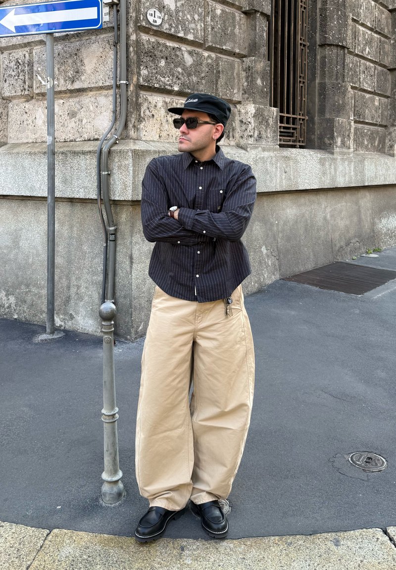 Chemise à rayures noires à manches longues, pantalons beige à jambes larges, chaussures noires et casquette. Se tient contre un mur en pierre, les bras croisés.