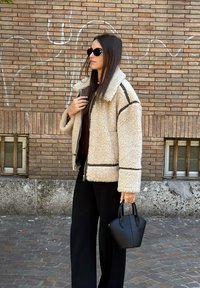 Jachetă teddy bej cu dungi contrastante maro, închidere cu fermoar și guler. Geantă neagră cu mânere scurte. Pantaloni negri. Fundal cu zid de cărămidă.