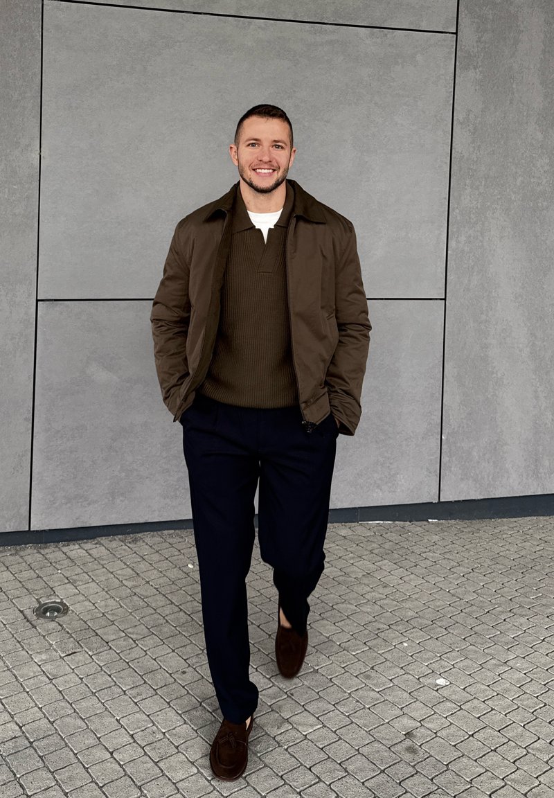 Smiling man walking on tiled pavement wearing brown jacket, dark jumper, navy trousers, and brown loafers against grey panel wall background.