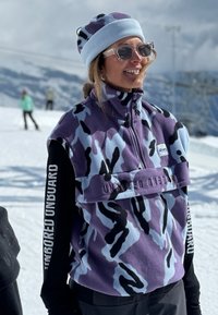 Mujer sonriente con gafas de sol, gorro estampado y chaleco polar camuflado morado con el texto "UNBORED", de pie en una pista de esquí nevada.