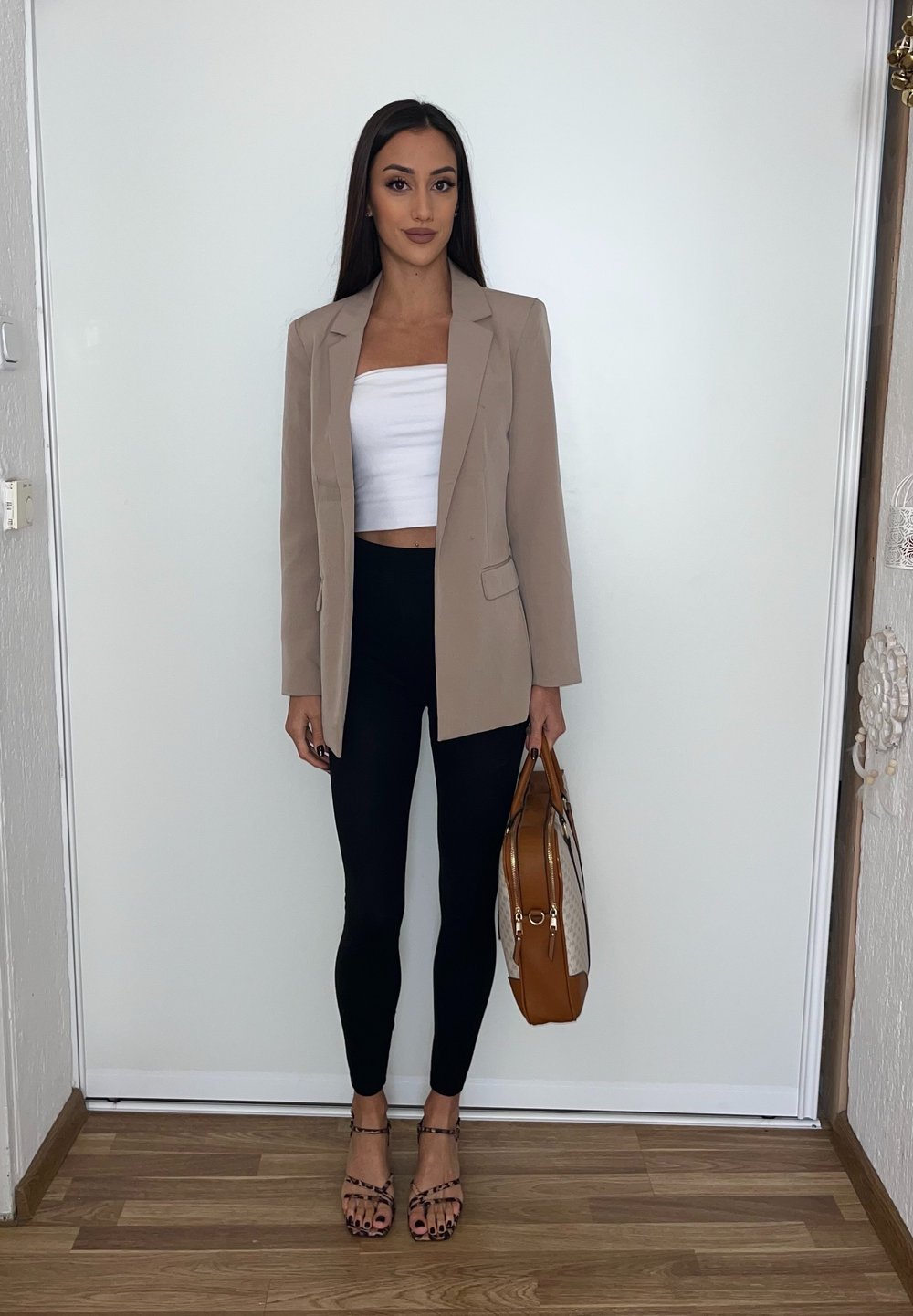 Béžový blejzer cez biely crop top, čierne legíny a sandály na opätku. Drží hnedú kabelku so vzorovým akcentom. Stojí na drevenej podlahe.