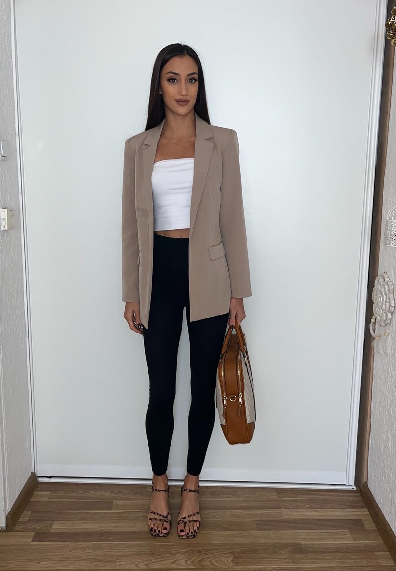 Beige blazer over a white crop top, black leggings, and strappy sandals. Holds a brown handbag with a patterned accent. Standing on wooden flooring.