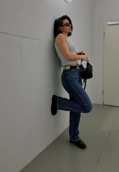 Una persona se apoya contra una pared gris, vistiendo una camiseta sin mangas gris claro, jeans azules, zapatos negros y gafas de sol, sosteniendo un pequeño bolso negro.