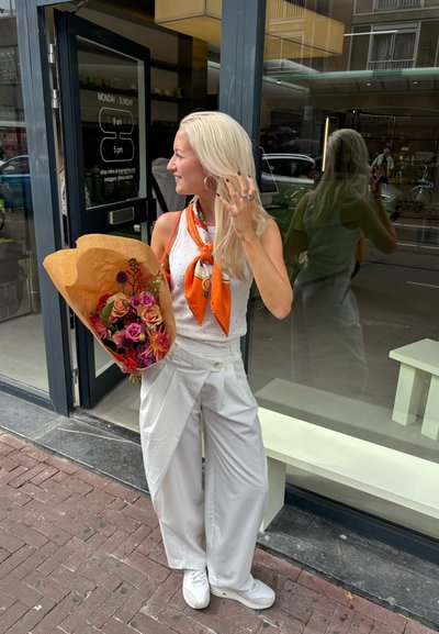 Una persona con una blusa blanca y pantalones de pierna ancha sostiene un ramo de flores envuelto en papel marrón cerca de una tienda con puertas de cristal.