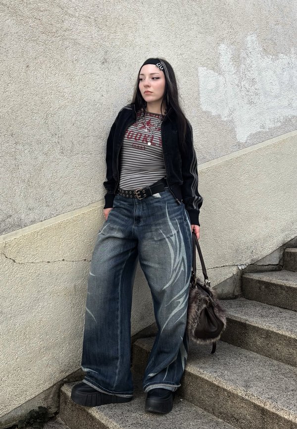 Giovane donna in piedi su gradini di pietra, indossa jeans a gamba larga con disegni bianchi, maglietta a righe, giacca nera, scarpe con plateau e tiene una borsa rifinita in pelliccia.