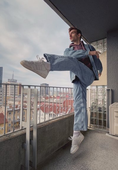 Chaqueta de mezclilla sobre una sudadera morada, jeans de pierna ancha azul claro y zapatillas blancas; persona realizando una patada alta en la barandilla de un balcón.
