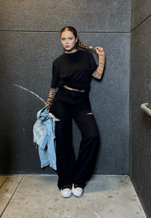 Top negro de gran tamaño con cortes laterales, pantalones negros de pierna ancha, chaqueta de mezclilla y zapatillas blancas. La modelo tiene el cabello largo trenzado.