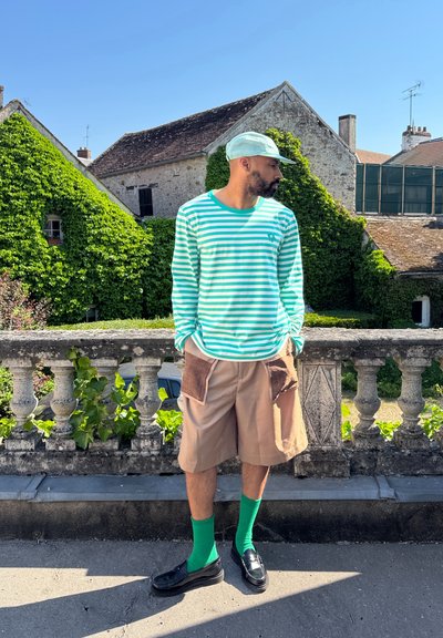 Camisa de manga larga a rayas en menta y blanco, pantalones cortos beige con bolsillos de parche, medias verdes hasta la rodilla y mocasines negros. Entorno al aire libre, con vegetación de fondo.