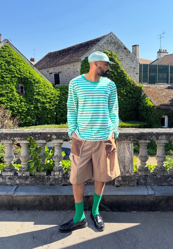 Camicia a maniche lunghe a righe in menta e bianco, pantaloniorti beige con tasche a toppa, calzettoni verdi fino al ginocchio e mocassini neri. Ambientazione all'aperto, verdure sullo sfondo.