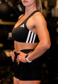 Fit woman wearing black Adidas sportswear and a white smartwatch stands with hands on hips in a gym setting.