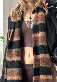 Gestrickter Schal in Schwarz-, Braun- und Grüntönen mit Wollstruktur. Getragen über einem braunen, taillierten Top mit V-Ausschnitt. Geschichtet mit einer schwarzen Lederjacke.