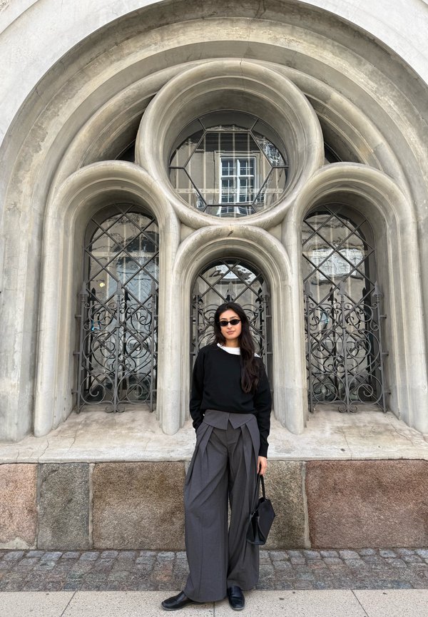 Woman in black sweater and wide gray pants stands holding handbag in front of a building with three rounded arched windows and ornate iron grilles.