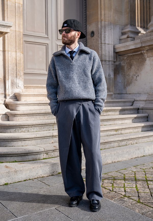 Homme barbu portant une casquette noire, des lunettes de soleil, un pull en maille gris, un pantalon large gris et des chaussures noires, debout à l'extérieur près de marches en pierre.