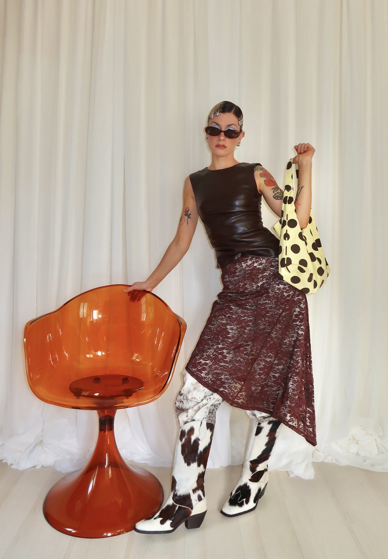 Femme portant un haut sans manches en cuir noir, une jupe en dentelle bordeaux, des bottes à motif vache, tenant un sac jaune à pois à côté d'une chaise moderne orange.