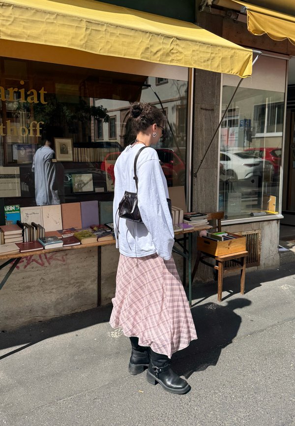 Femme avec un chignon et des lunettes de soleil feuilletant des livres sur une table extérieure sous un auvent jaune devant une vitrine par une journée ensoleillée.